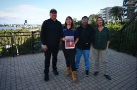 Four officials stand on a paved balcony overlooking Jackson Bay harbor on a sunny day; from left to right, a man in a black shirt and cap with a mustache, a woman in a blue blouse holding a red document, a man in a gray shirt and sunglasses, and a woman in a green blouse, with modern apartments, palm trees, and water visible in the background.