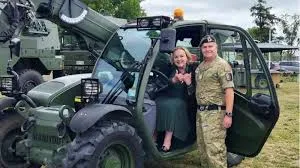 Defence Minister Judith Collins, in a green dress, and an army officer in camouflage uniform stand smiling with thumbs up inside the open cab of a green military telehandler at Burnham Military Camp, with additional military vehicles and equipment visible in the background under overcast skies.
