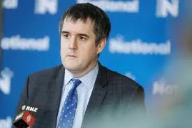 Transport Minister Chris Bishop stands at a podium during a press conference, wearing a grey suit, white shirt, and blue dotted tie with an RNZ lapel pin, against a blue backdrop featuring National Party branding.