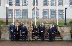 A group of 16 men and women, including military personnel in camouflage uniforms and berets alongside civilians in business suits, pose formally on stone steps in front of a large white stone building with blue windows, flagpoles, and outdoor lamps in Edinburgh.