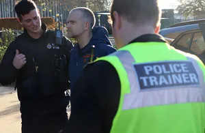 A police trainer in a yellow high-visibility vest stands facing two apprentice officers in black tactical vests during an outdoor training exercise. The trainer observes the apprentices near industrial fencing and parked vehicles under daylight conditions, with one apprentice gesturing and the other listening attentively.
