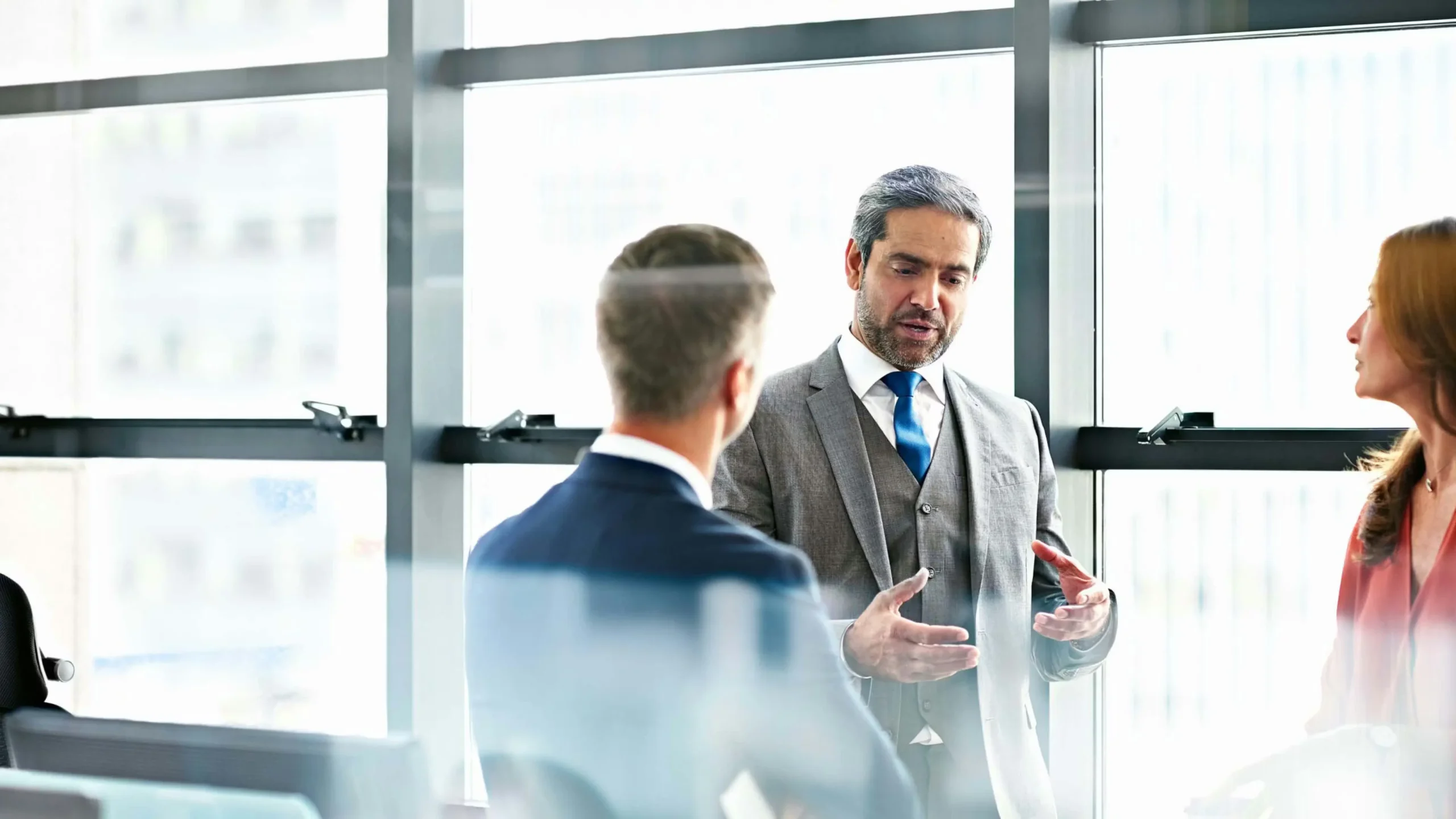 Three professionals in formal business attire engaged in a serious discussion inside a modern glass-walled office meeting room, with natural light from large windows.