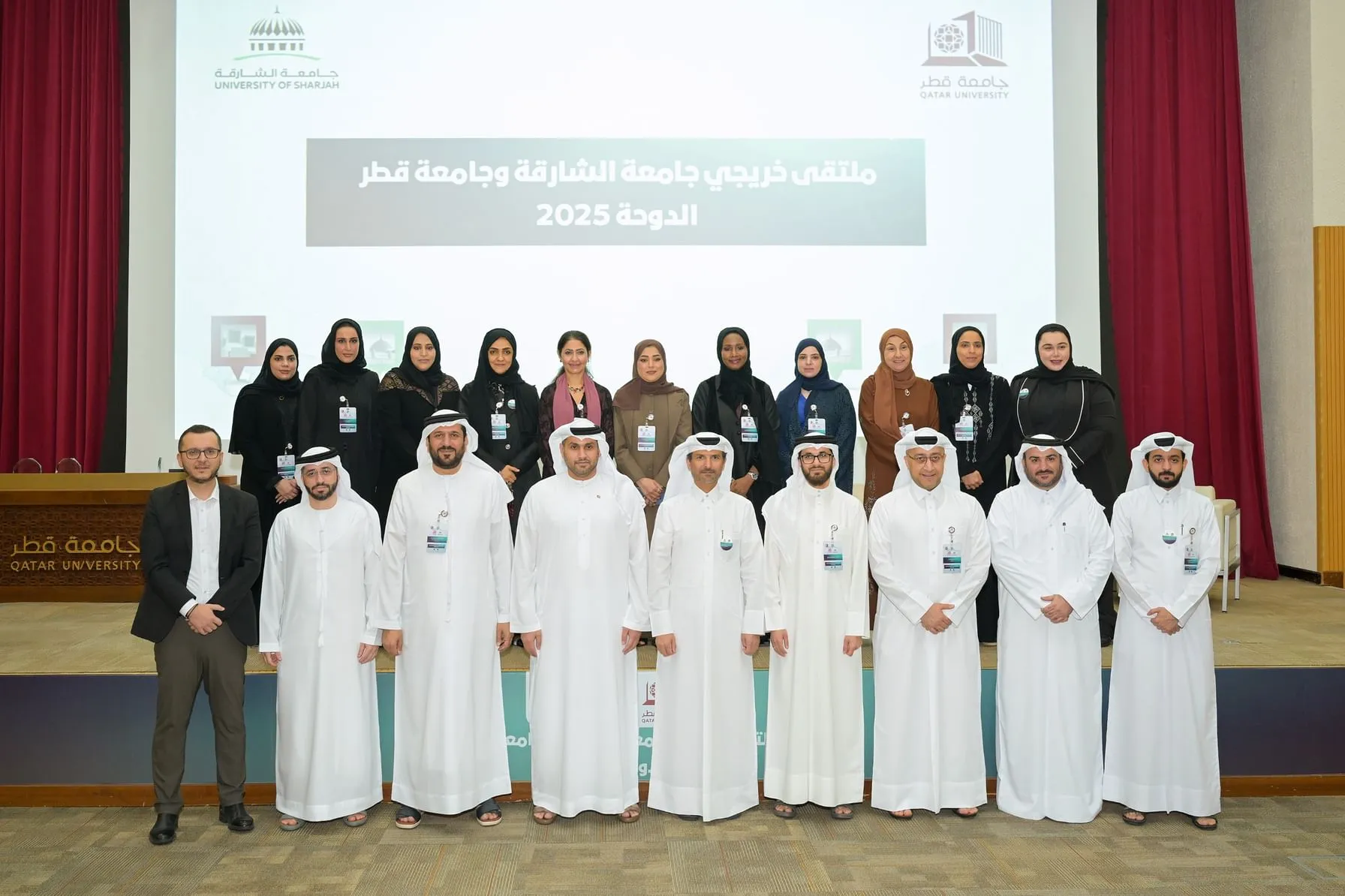 Group photo of the University of Sharjah Alumni Association Executive Committee members in Qatar, standing on a stage in front of a large screen displaying the title "University of Sharjah Alumni Association Qatar Chapter 2025" in Arabic and English, with logos of University of Sharjah and Qatar University. The group includes eight women in hijabs and abayas wearing name badges, and eight men in traditional white dishdashas and ghutras or Western suits, all positioned in two rows against a red curtain backdrop in an auditorium setting.