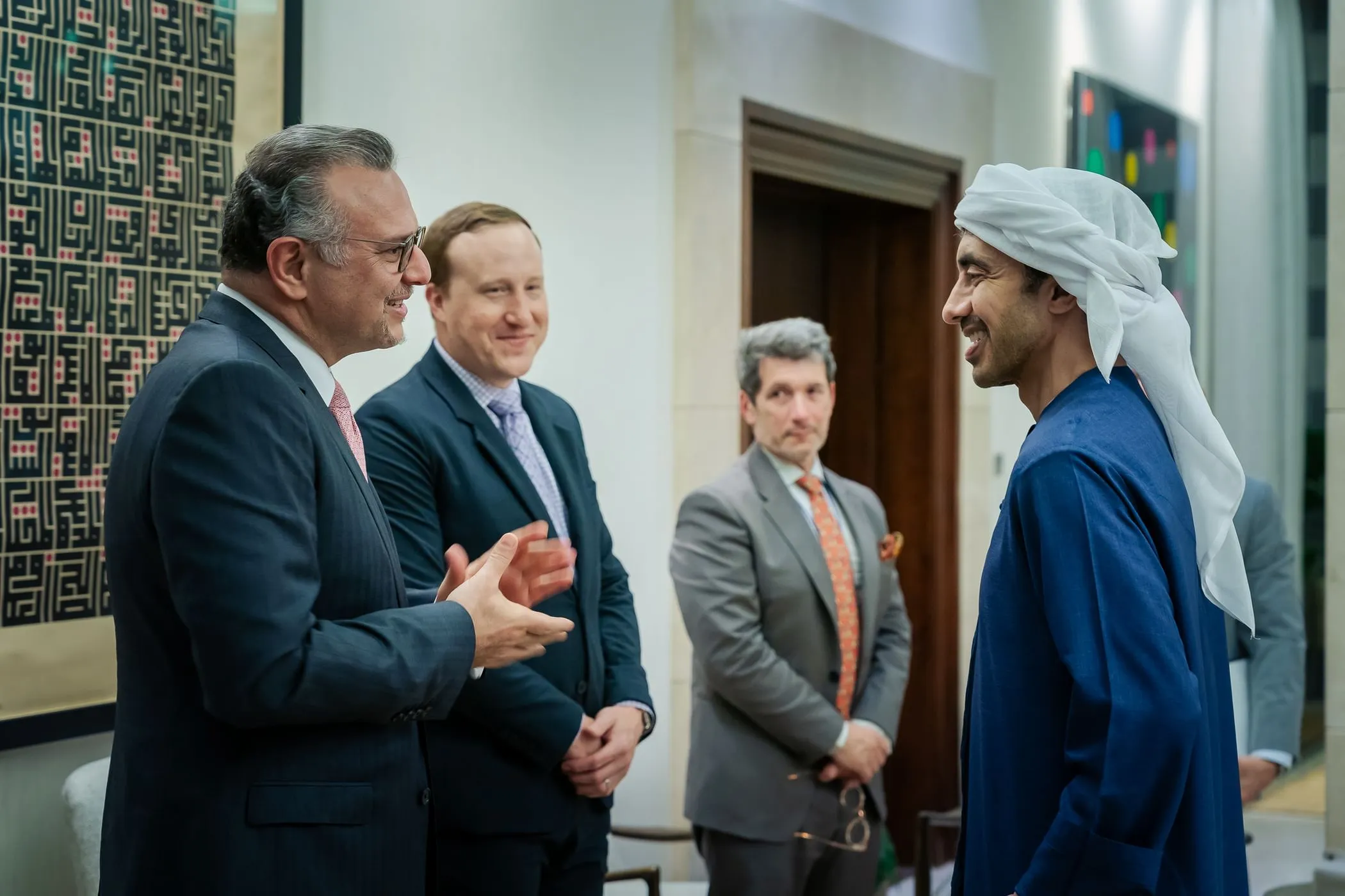In a formal meeting room in Abu Dhabi with geometric patterned wall art and a wooden door in the background, H.H. Sheikh Abdullah bin Zayed Al Nahyan, UAE Minister of Foreign Affairs, wearing a dark suit, white shirt, red tie, and glasses, gestures with his hands while conversing. Beside him stands a man in a blue suit, light blue shirt, and purple tie. Massad Boulos, US Senior Advisor for African and Arab Affairs, dressed in a blue dishdasha and white ghutra, smiles attentively. Two other men in gray suits stand nearby, observing the discussion.