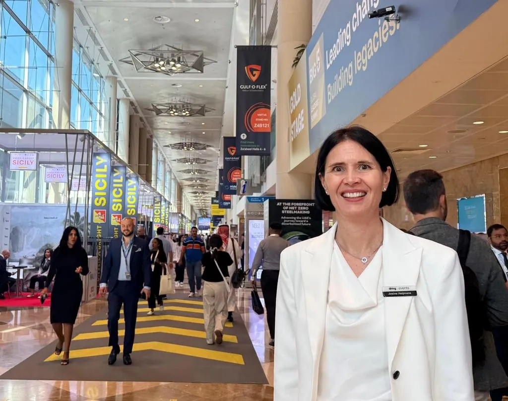 A wide-angle view of the bustling main hall at the Big 5 Global 2025 construction exhibition in Dubai World Trade Centre, featuring international exhibitors in glass display booths, yellow directional floor markings, overhead lighting, and banners for Gulf Flex and Gold Building Stage. In the foreground, Josine Heijmans, Senior Vice President of Construction at dmg events, smiles confidently while wearing a white draped blouse, name badge, and carrying a black bag, standing among diverse professionals in business attire.
