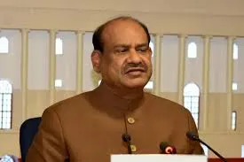 Lok Sabha Speaker Om Birla, dressed in a beige kurta, speaks at a podium during a Constitution Day event at Manipal University in Jaipur, with microphones and a blurred audience in the background.
