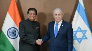Indian Commerce Minister Piyush Goyal, wearing a traditional maroon kurta, shakes hands with Israeli Prime Minister Benjamin Netanyahu, dressed in a blue suit and tie, in front of the Indian and Israeli national flags against a brown backdrop.