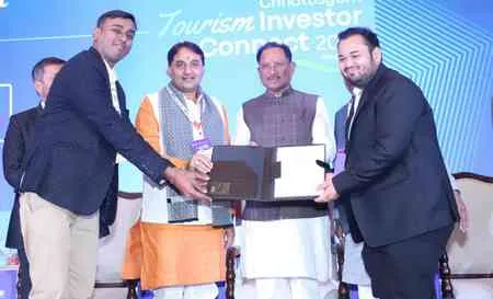 Four men in formal Indian attire stand on a stage during the Tourism Investor Connect 2025 event. The central two men, one wearing an orange kurta and white shawl and the other a cream kurta, shake hands while exchanging a laptop and a white folder. The other two men, in navy suits, stand beside them holding lanyards. The blue backdrop features the event logo and name.