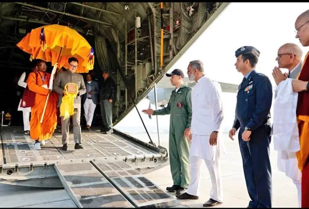 Union Minister Kiren Rijiju, dressed in a grey suit, holds a yellow-wrapped box containing sacred Buddha relics and walks down the ramp of an Indian Air Force aircraft at Palam Airport. He is accompanied by orange-robed Buddhist monks carrying orange umbrellas and staff, and greeted by uniformed air force officers in green and blue uniforms, white-clad officials, and additional monks in maroon robes standing on the tarmac.