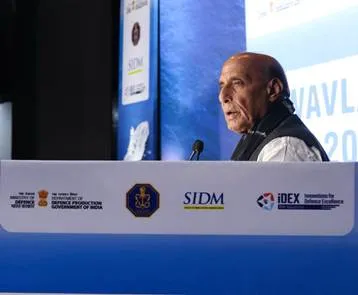 Raksha Mantri Rajnath Singh, an elderly man with glasses and white beard wearing a light kurta and shawl, speaks into a microphone at a podium during the Swavlamban 2025 seminar, with a blue backdrop displaying the event name and logos of Department of Defence Production, Government of India, SIDM, and iDEX.