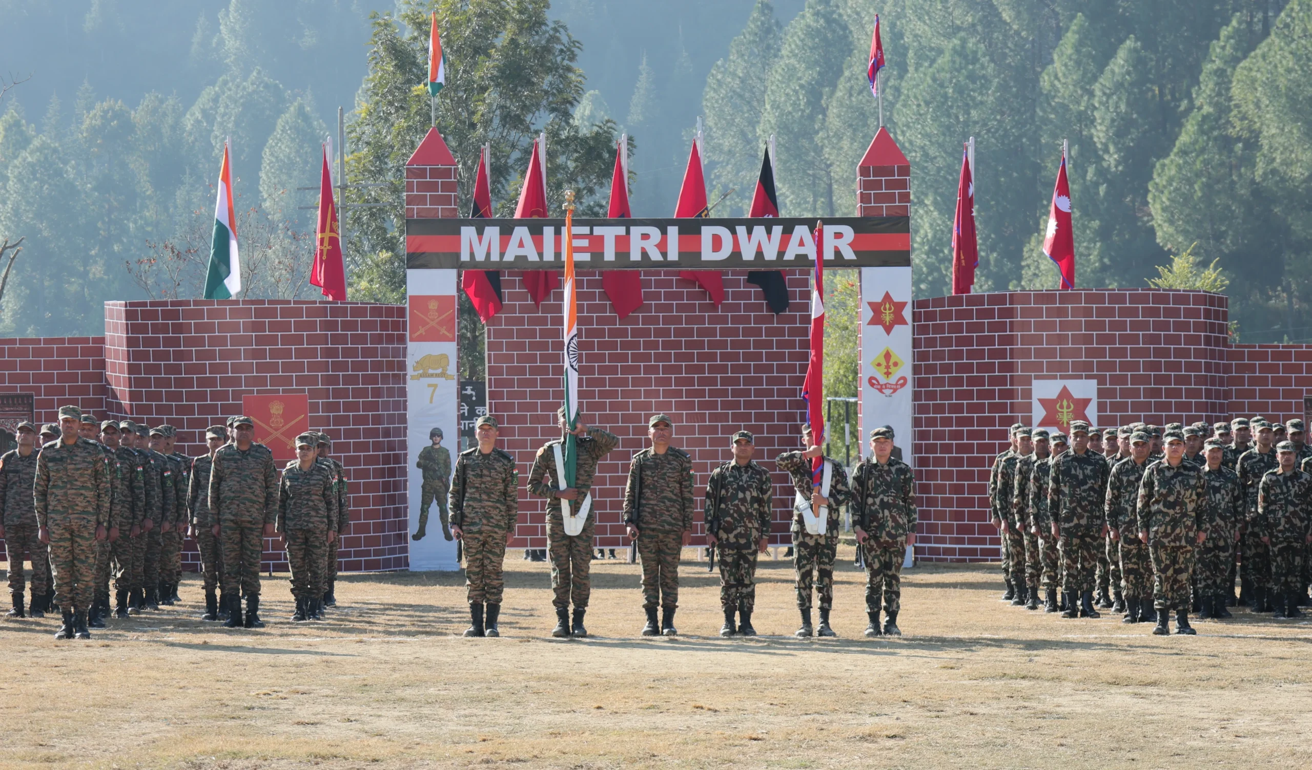 Indian and Nepali soldiers in camouflage uniforms stand in formation during the inauguration ceremony of Exercise Suryakiran XIX at the Maitri Dwar gate in Pithoragarh, Uttarakhand, with Indian and Nepali national flags, military emblems, and a brick archway backdrop against a mountainous forested background.