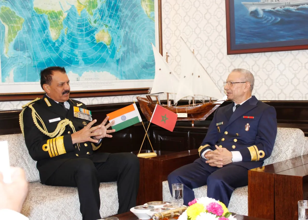 Two naval officers seated on a couch in a formal room with a world map and maritime painting on the walls. The officer on the left, in an Indian Navy uniform with epaulettes and medals, holds an Indian flag while gesturing. The officer on the right, in a Moroccan Navy uniform, sits attentively. Small Indian and Moroccan flags stand on a table between them, alongside a model wooden sailing ship, a brass telescope, glasses of water, and a vase of pink and yellow flowers.