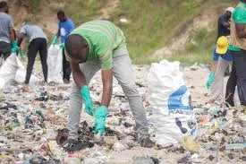 A group of volunteers wearing green t-shirts, gray pants, and green gloves bend down to pick up plastic waste from a sandy riverbank littered with trash, filling large white trash bags labeled 'PLASTIC' as part of a community cleanup initiative.
