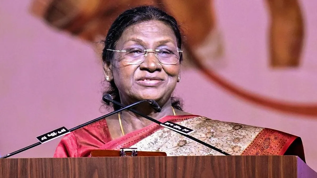 President Droupadi Murmu, wearing a red and gold saree and glasses, speaks at a podium with a microphone during the National Constitution Day function, with pink and orange decorative backdrops and a wooden lectern in the Central Hall of Samvidhan Sadan.