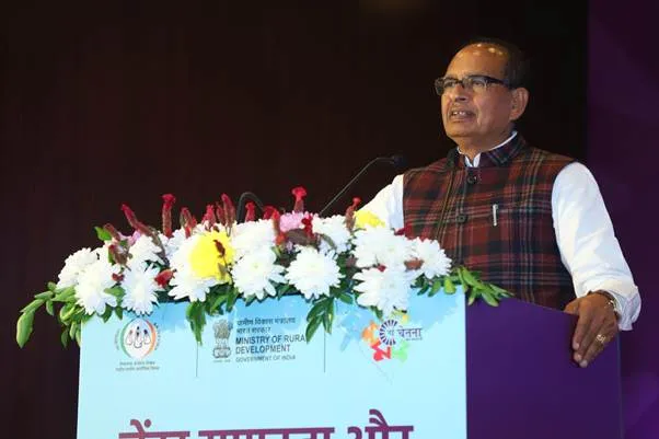Union Minister Shivraj Singh Chouhan, wearing a maroon plaid vest over a white kurta, stands at a podium adorned with white and yellow flowers, speaking into a microphone during the inauguration event. The podium displays logos of the Ministry of Rural Development, Government of India, and a blue banner with Hindi text reading 'नई चेतना अभियान' against a purple backdrop at Pravasi Bharatiya Kendra in New Delhi.