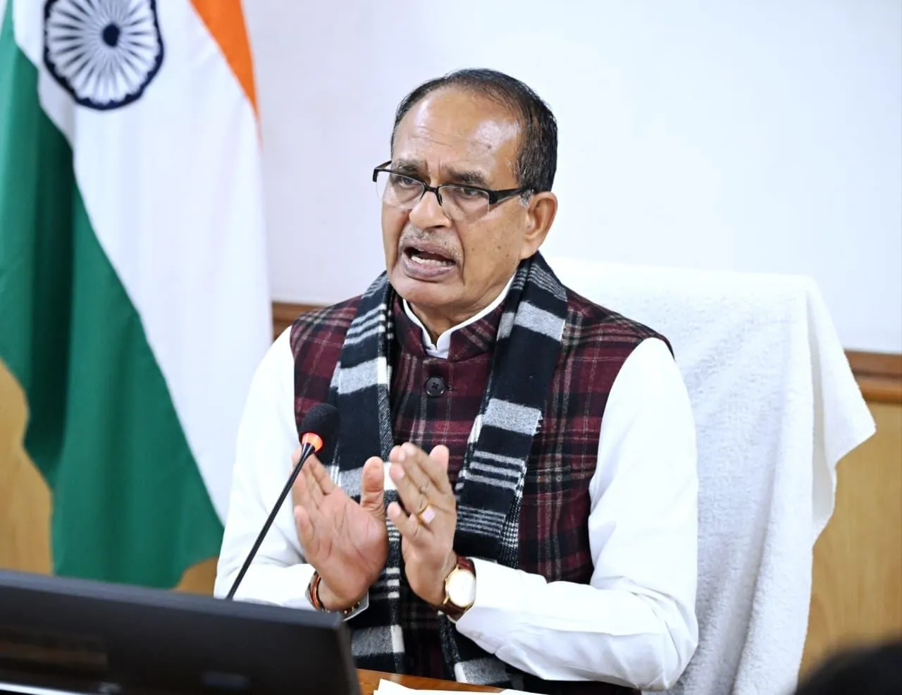 Union Minister Shivraj Singh Chouhan, wearing a maroon vest with black scarf and white shirt, speaks into a microphone with hands clasped at a podium during a video conference, with Indian national flags and a laptop in the background.