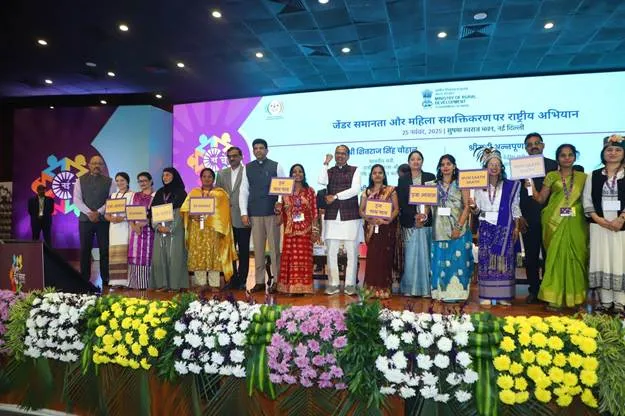Clear, descriptive, factual (no opinions). Must fully describe the visual content for screen readers. | A diverse group of Indian government officials and women leaders, including men in suits and kurtas and women in traditional sarees and salwar kameez, standing on a decorated stage holding framed certificates or placards. The backdrop features a purple and white logo for the Nayi Chetna 4.0 campaign with Hindi and English text on gender equality and women empowerment, floral garlands in yellow, white, and pink below, and the event location at Sushma Swaraj Bhawan in New Delhi.