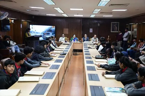 A diverse group of students in school uniforms and officials in formal attire seated around a long wooden conference table in a paneled room with large windows and a ceiling fan. A projector screen at the front displays event visuals, while participants, including speakers at a podium, engage during the Curtain Raiser for India International Science Festival 2025 at CSIR-NIScPR, New Delhi.