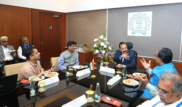 A group of eight Indian men in formal and traditional attire, including kurtas and suits, seated around a polished black conference table in a modern wood-paneled meeting room. They are engaged in discussion, with some gesturing and others eating from white plates with Indian dishes. The table features Indian national flags, glass water bottles, flower arrangements, and name placards. A wall-mounted banner displays a logo with interlocking gears and a lotus emblem, likely representing an Indian scientific institution.
