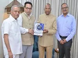 Four senior Indian officials standing side by side in a conference room, dressed in traditional attire: an elderly man in a white half-sleeve shirt and trousers on the left, a man in a beige kurta with vest and glasses next to him, an elderly man in a cream kurta pajama holding a blue-covered booklet with white text and circular logo in the center, and a man in a blue shirt and trousers with glasses on the right. The backdrop includes beige curtains and wooden chairs, indicative of an official event in Mumbai.