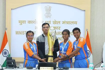 Union Minister Dr. Mansukh Mandaviya, wearing a gray shirt, stands in the center holding a golden trophy with four members of the Indian women's blind cricket team dressed in orange jerseys. The group poses in front of a banner reading "Ministry of Youth Affairs & Sports" in Hindi and English, with Indian flags on either side. The trophy base is inscribed "Dr. Mansukh Mandaviya.