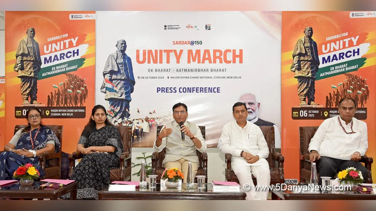 Five Indian dignitaries seated at a press conference table in front of orange banners promoting the Sardar@150 Unity March event on October 6, 2025, at Major Dhyan Chand National Stadium in New Delhi. The central male speaker in a beige kurta holds a microphone, flanked by two women in colorful saris and two men in white attire, with Indian flags, floral arrangements, water glasses, and documents on the wooden table; event logos and credit to 5DarryaNews visible.