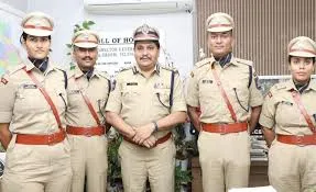 Five Indian police officers, including two women wearing caps and khaki uniforms with red sashes and shoulder epaulettes, stand in a row with hands clasped in front, positioned indoors in front of a wall-mounted air conditioning unit, a sign reading "HALL OF HONOUR," and potted green plants on either side.