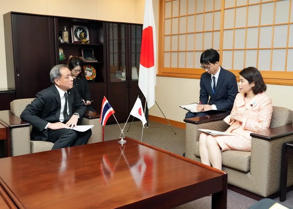 Dr. Ayano Kunimitsu, Japan's State Minister for Foreign Affairs, seated across a wooden table from H.E. Witchu Vejjajiva, Ambassador of Thailand to Japan, in a traditional Japanese office with national flags of Japan and Thailand displayed prominently in the background. Both are dressed in formal attire, holding documents, with bookshelves and decorative items visible.