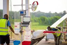 Two workers in yellow safety vests prepare a white fixed-wing drone with red accents at an outdoor launch station in a grassy field in Africa. The setup includes a tall metal mast with a control computer, monitor, battery pack, and a red safety light; a silver reflector nearby; and storage boxes on the ground.