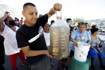 A young man in a black shirt holds up a large plastic jug filled with murky water in front of a crowd of diverse people, including women and men wearing face masks, gathered outdoors near green water containers and parked cars under a sunny sky.