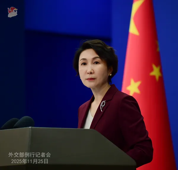 Chinese Foreign Ministry Spokesperson Mao Ning stands at a podium during a regular press conference on November 26, 2025, wearing a maroon blazer over a white blouse with a brooch, speaking into microphones with a serious expression; a Chinese national flag is positioned to her right against a blue backdrop.