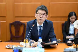 A man wearing glasses and a dark suit speaks into a microphone at a formal conference table during an official meeting, with a nameplate reading "H.E. Aun Pornmoniroth" in front of him and another participant visible in the background.