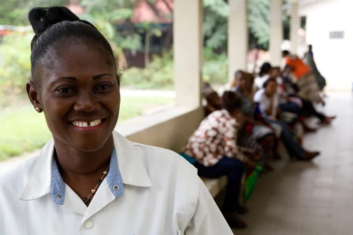 A smiling Black woman in a white collared shirt with blue collar trim and black bead necklace, her hair in a black bun, stands in the foreground at an outdoor clinic covered by a porch with green vegetation in the background; several people of various ages sit on benches behind her, waiting in a tropical setting.
