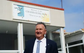 A man in a dark suit and blue tie stands smiling in front of a building facade featuring a large sign that reads "Welcome to Chatham Islands" with a sun emblem and a stylized wave or dolphin logo, under a clear blue sky.