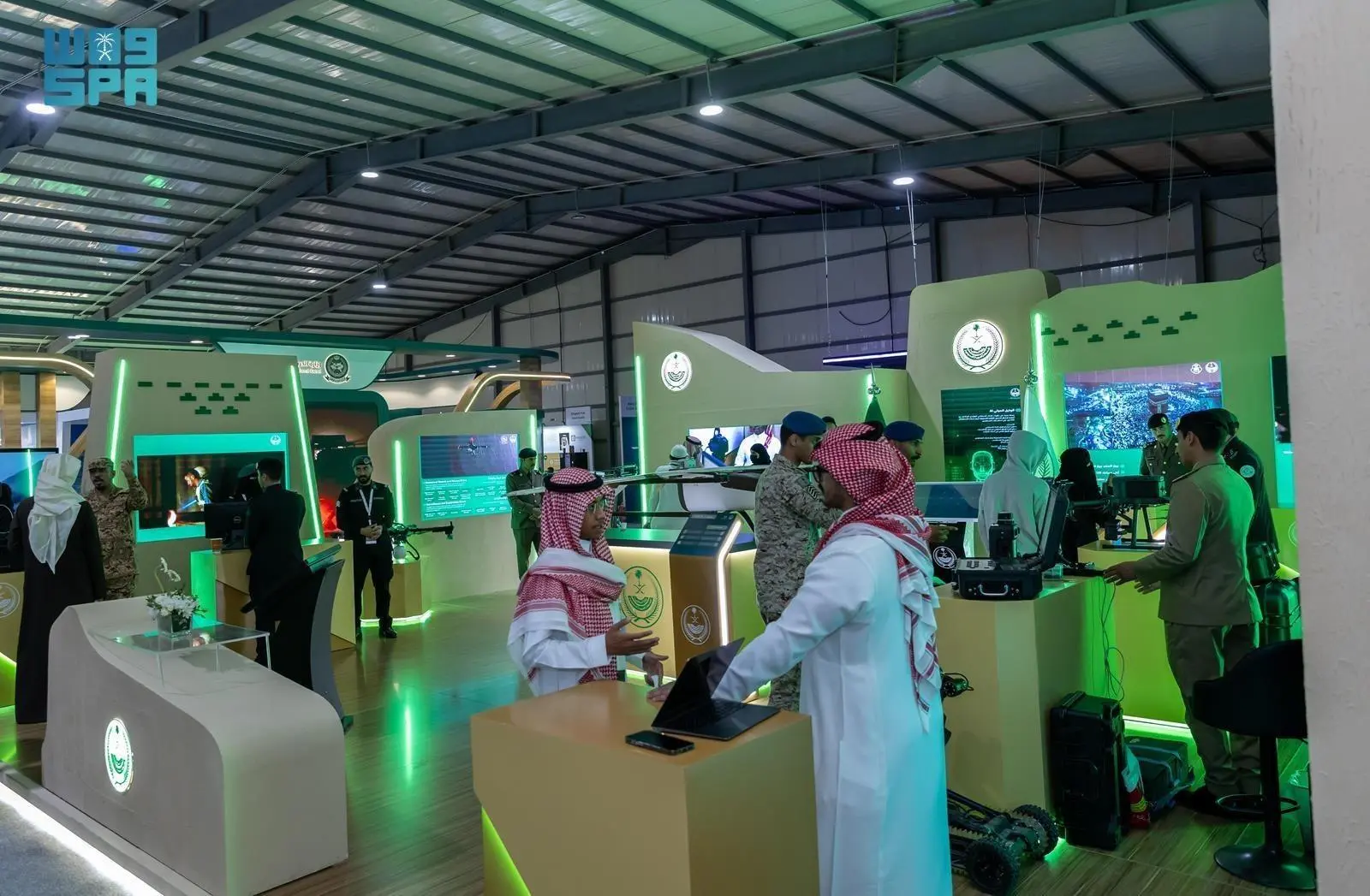 A vibrant exhibition hall at the Sand and Fun 2025 General Aviation Airshow in Riyadh, featuring the Saudi Ministry of Interior pavilion with curved white and green booths displaying AI technology screens, early warning platforms, and interactive displays. Officials in traditional Saudi attire, military uniforms, and business suits engage in discussions near laptops and gadgets, including a small robot and drone equipment, under bright overhead lighting and green accent illumination.