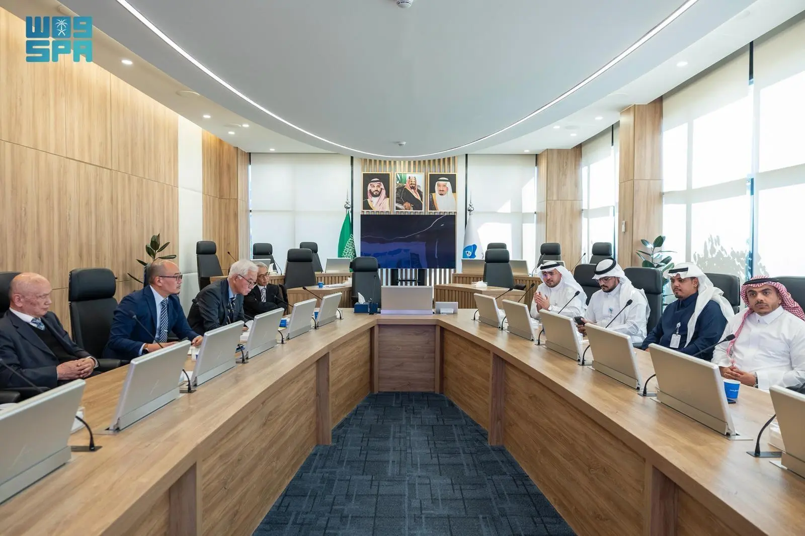 A formal meeting in a modern conference room at the Saudi Water Authority headquarters in Riyadh, featuring a U-shaped wooden table with laptops and documents. Seated attendees include Saudi officials in traditional white thobes and ghutras, and international delegates in Western business suits. Portraits of Saudi leaders adorn the wall behind a large screen, flanked by Saudi and partner country flags including Japan, Portugal, and Korea.