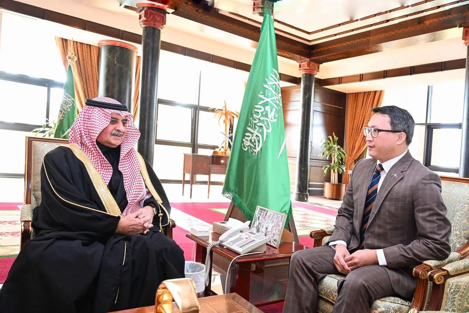 Saudi Prince Fahd bin Sultan bin Abdulaziz, wearing traditional white thobe, red and white checkered headscarf and black agal, sits in an ornate wooden armchair in a grand room adorned with Saudi flags. He is engaged in conversation with Rommel Romato, the Filipino Consul General, dressed in a gray suit, blue striped tie, and white shirt with a Philippine flag pin, seated opposite across a wooden table with a white telephone and decorative vase. The room features marble columns, red carpet, large windows, potted plants, and framed photos.