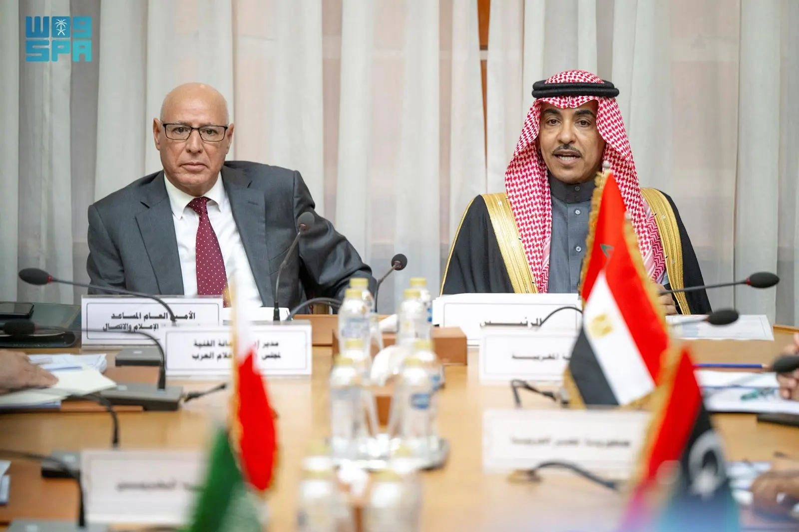 Saudi Minister of Media Salman Al-Dossary, wearing a red-and-white keffiyeh and black bisht, seated at a conference table with Egyptian Prime Minister Mostafa Madbouly, dressed in a gray suit and red tie, during a bilateral meeting in Cairo. UAE, Egyptian, and Saudi flags are positioned on the table, with microphones, water bottles, and documents visible in front of them, against a backdrop of white curtains and the WAM-SPA logo in the corner.