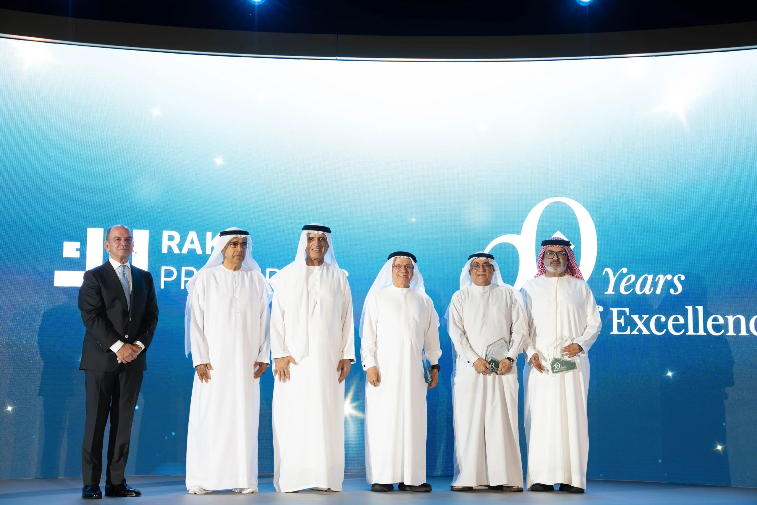 Six men standing on a stage during a formal event, with five wearing traditional white Emirati dishdashas and ghutras, and one in a dark suit. The man in the suit holds a green plaque, while two others hold trophies. The backdrop features the RAK Properties logo, "20 Years of Excellence" text, and blue lighting with spotlights.