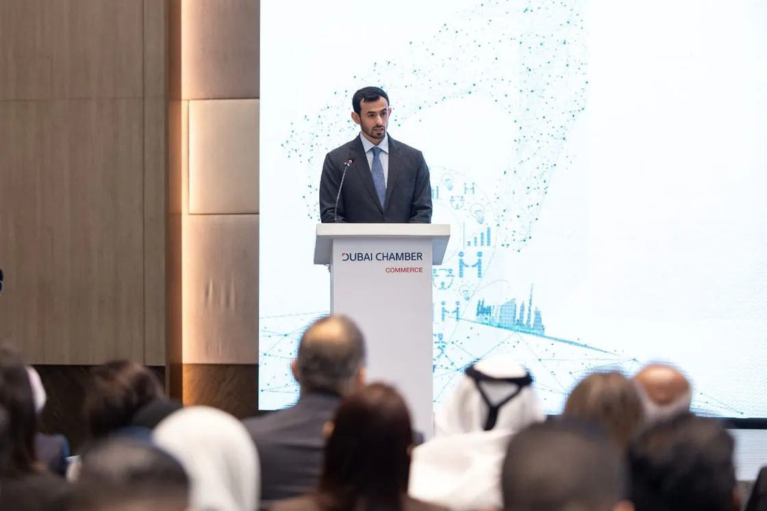 Salem AlShamsi, Executive Vice President of International Relations at Dubai Chambers, speaks at a podium marked with the Dubai Chamber of Commerce logo during a trade mission event in Kuala Lumpur. The backdrop features digital graphics of networks, charts, and Dubai skyline silhouettes. Audience members in business attire, including some wearing traditional headgear, are seated and listening attentively.