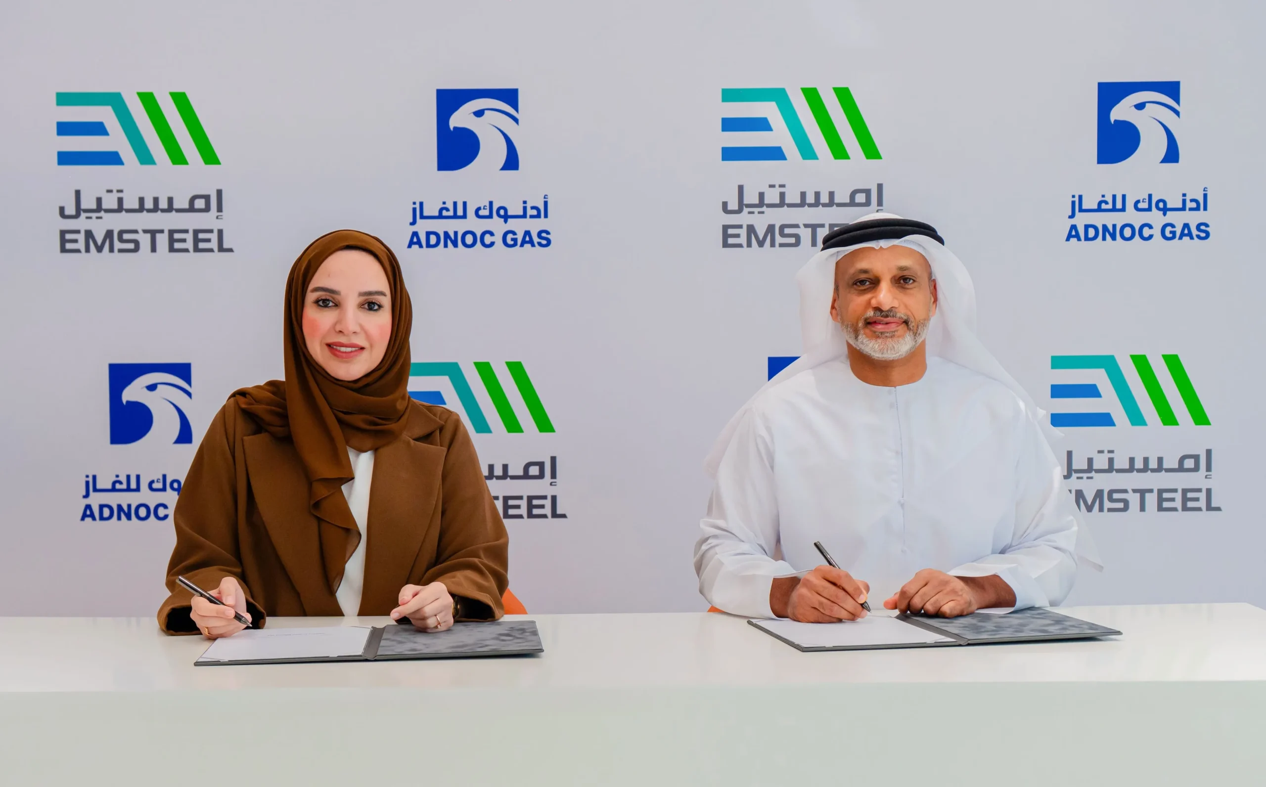 Two individuals seated at a conference table signing documents on a white surface. The woman on the left wears a brown hijab and blazer, holds a pen, and has documents in front of her. The man on the right wears a white traditional dishdasha and ghutra, holds a pen, and has a clipboard. The backdrop features logos of EMSTEEL and ADNOC Gas in blue and green colors with Arabic and English text.