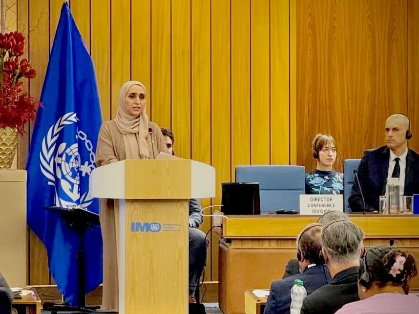 Eng Hessa Al Malek, a woman wearing a beige hijab and coat, speaks at a podium with the IMO logo during the 34th session of the International Maritime Organisation Assembly in London, with UN and IMO flags in the background, and delegates seated at a panel including a bald man with headphones and a woman with short hair wearing a floral blouse.