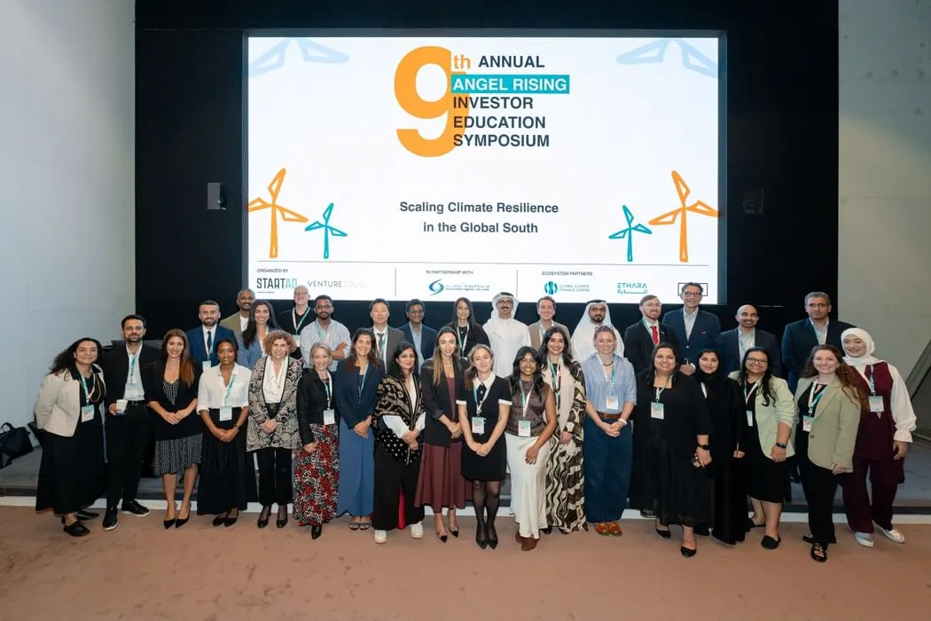 Group photo of diverse participants, including men and women in business attire and traditional garments, standing on a stage in front of a large screen displaying "9th Annual Angel Rising Investor Education Symposium" with the theme "Scaling Climate Resilience in the Global South," organized by startAD and VentureSouq in partnership with EAD, at Abu Dhabi Global Market.