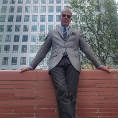A middle-aged man with a bald head wearing dark sunglasses, a light gray suit, light blue shirt, and pocket square, stands leaning against a red brick wall with arms crossed on the ledge, in front of a modern glass office building surrounded by green trees.