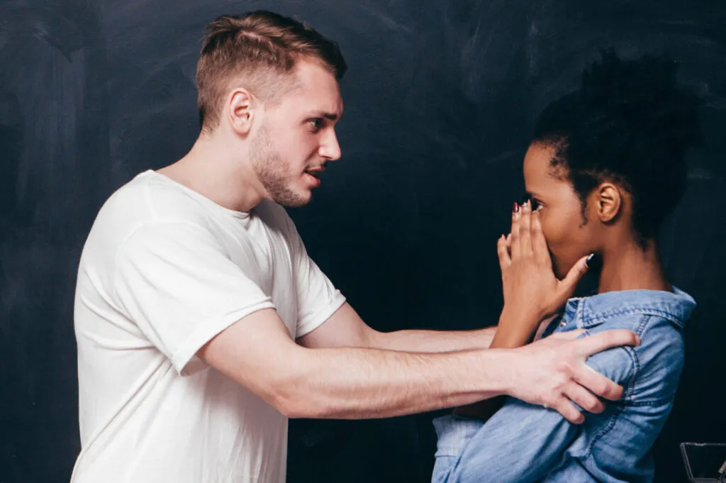 A young man with short blond hair and a white t-shirt stands facing a young Black woman with curly hair wearing a blue denim jacket, who covers her face with both hands in distress while he places a hand on her shoulder, set against a dark gray background.