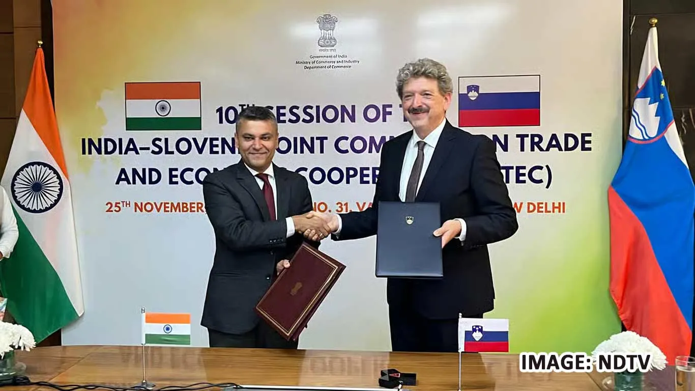 Two officials, Joint Secretary Saket Kumar from India and Director General Peter Japelj from Slovenia, shake hands after co-chairing the 10th Session of the India-Slovenia Joint Committee on Trade and Economic Cooperation (JCTEC) in New Delhi on 25 November, holding signed documents in front of Indian and Slovenian flags.