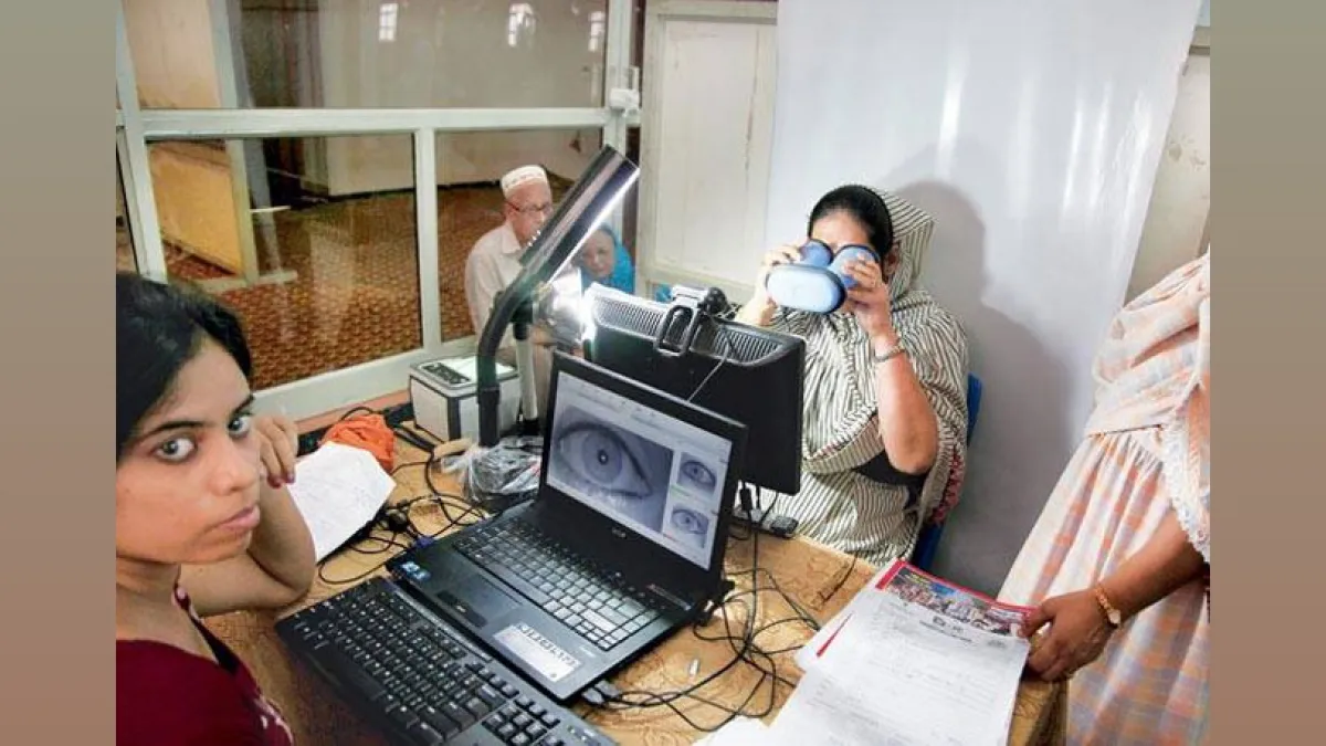 A woman in a striped headscarf undergoes iris scanning for Aadhaar biometric enrolment at a centre in India, while an operator monitors the eye image on a laptop screen.