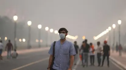A man wearing a light blue short-sleeved shirt, black cap, and N95 mask walks along a wide, empty road in Delhi, India, amid dense winter smog that reduces visibility. Streetlights glow in the hazy background, with a few pedestrians and cyclists visible in the fog-shrouded street, highlighting severe air pollution conditions.