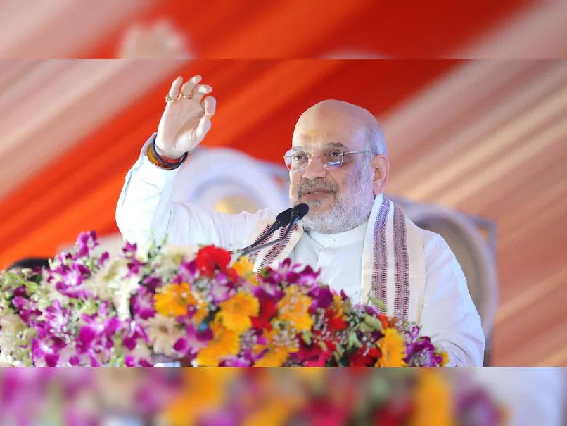 Union Home Minister Amit Shah waves to the crowd while addressing a public event from the podium in a white kurta and shawl.
