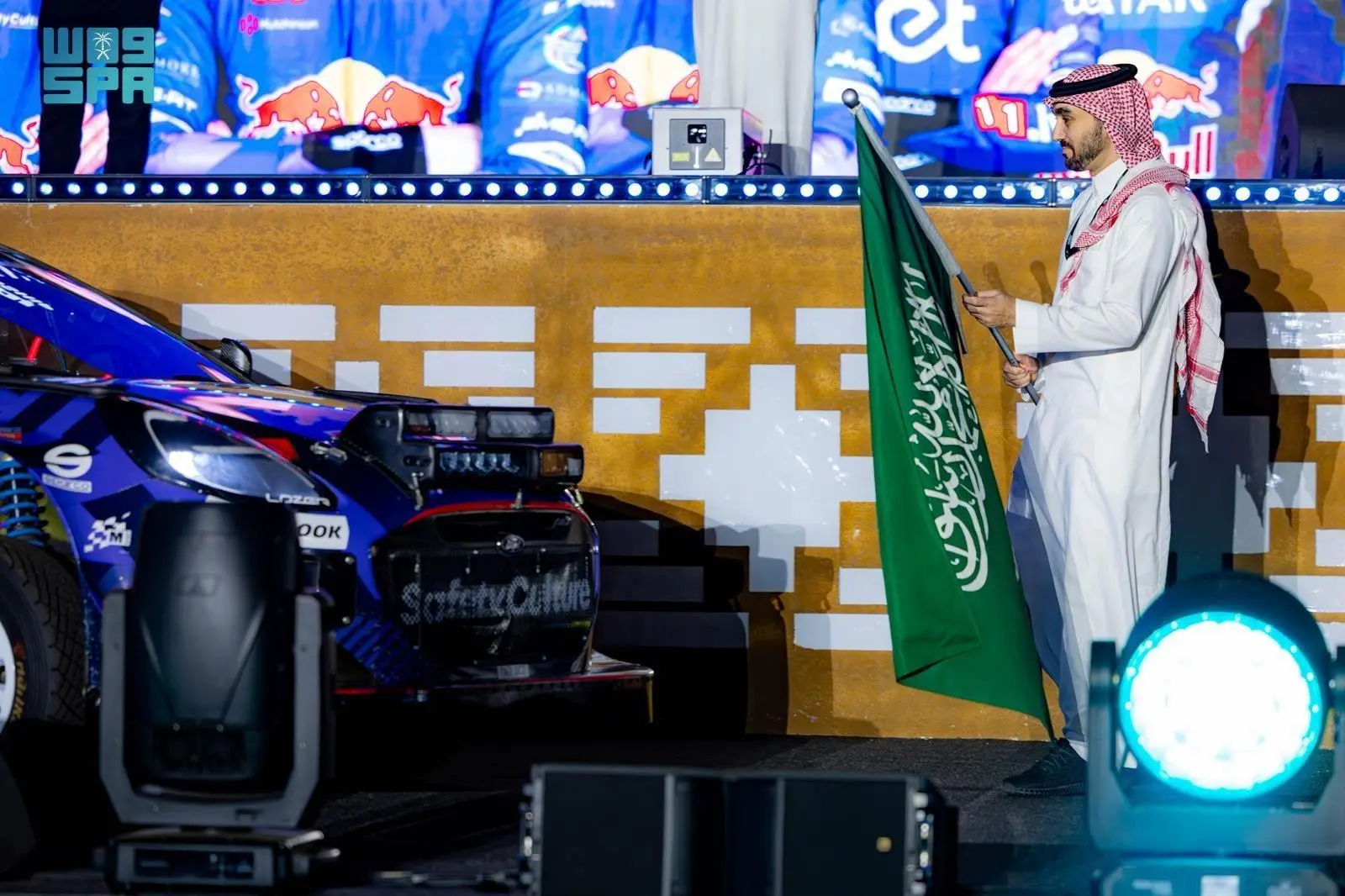 Prince Abdulaziz bin Turki Al Faisal holds the Saudi Arabian flag next to a Hyundai i20 WRC rally car during the ceremonial start of WRC Rally Saudi Arabia 2025 in Jeddah.